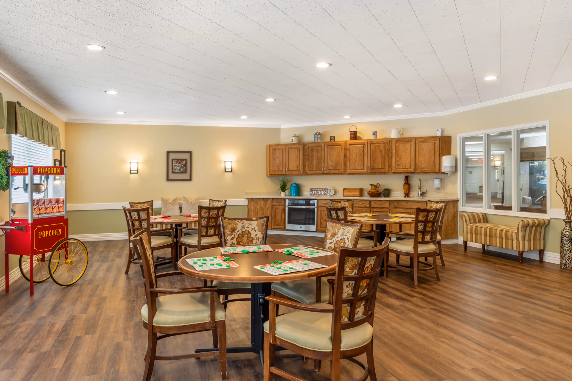 A spacious activity room with several round tables and chairs arranged for a game of bingo. The tables have bingo cards and markers on them. The room features wooden flooring, beige walls with green lower trim, and a popcorn machine on the left side. There is a kitchen area with wooden cabinets and a countertop along the back wall. A striped bench is placed near a window on the right side.