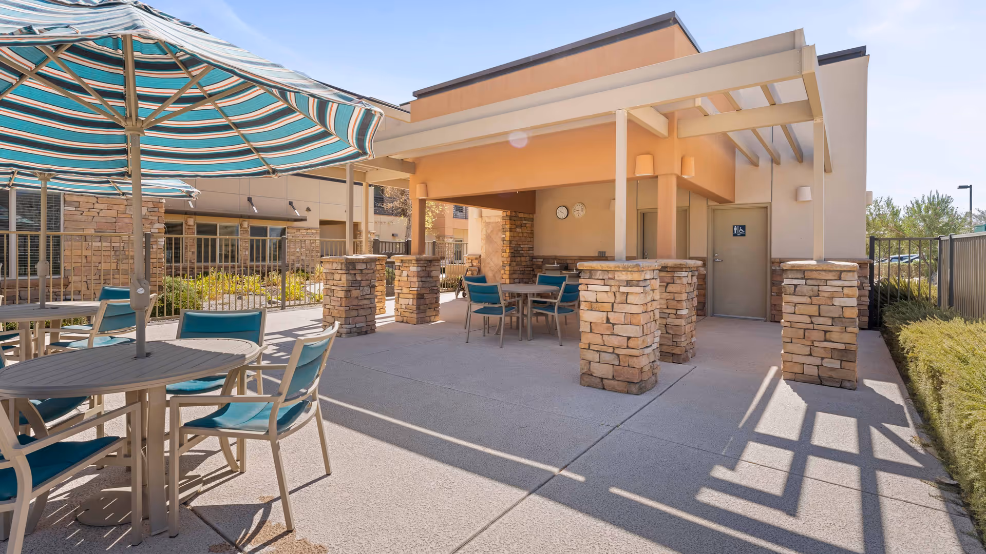 Outdoor patio area with several round tables and chairs with teal cushions, shaded by large striped umbrellas. The patio is adjacent to a building with stone pillars and a covered section. There is a door with a restroom sign and two clocks mounted on the wall under the covered area. The scene is bright and sunny with clear skies.
