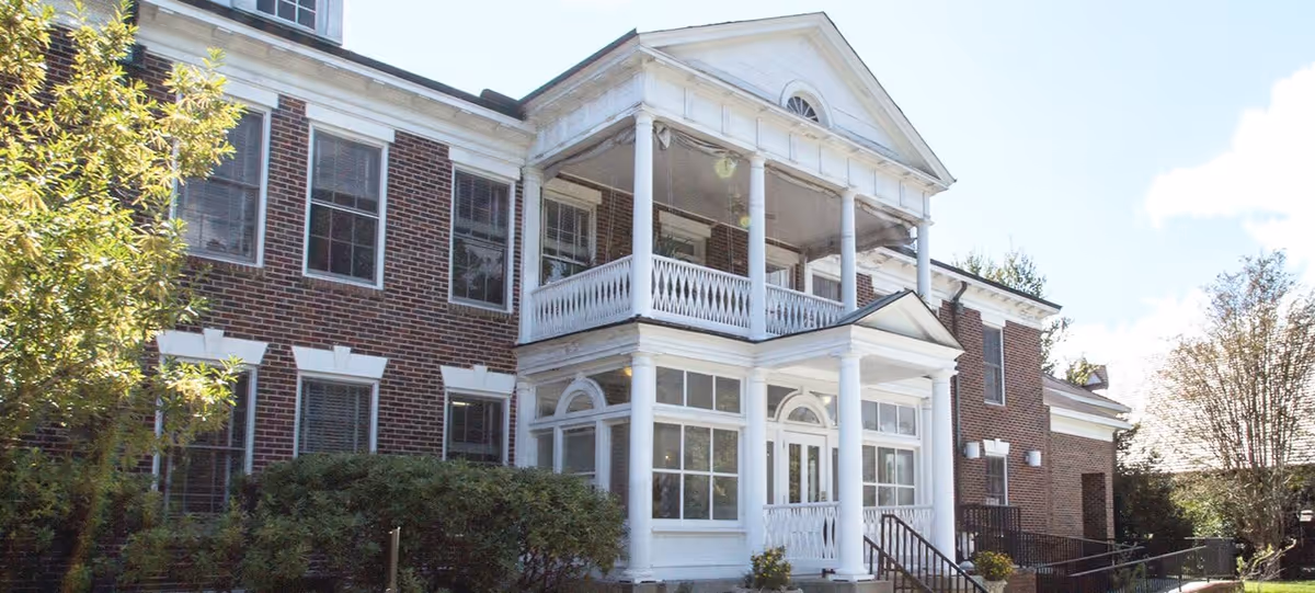 Exterior view of a two-story brick building with white columns and railings, featuring large windows and a covered porch area. There are bushes and trees around the building under a clear sky.