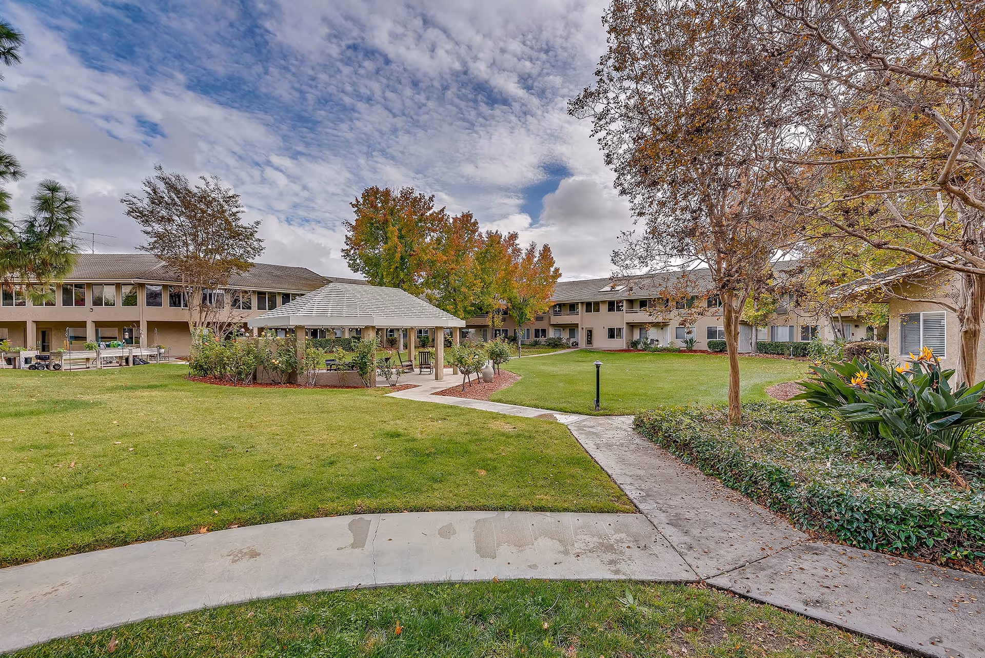 Outdoor courtyard area of Arcadia Place Senior Living featuring a green lawn, a covered gazebo with seating, surrounding two-story residential buildings, trees with autumn foliage, and a partly cloudy sky.