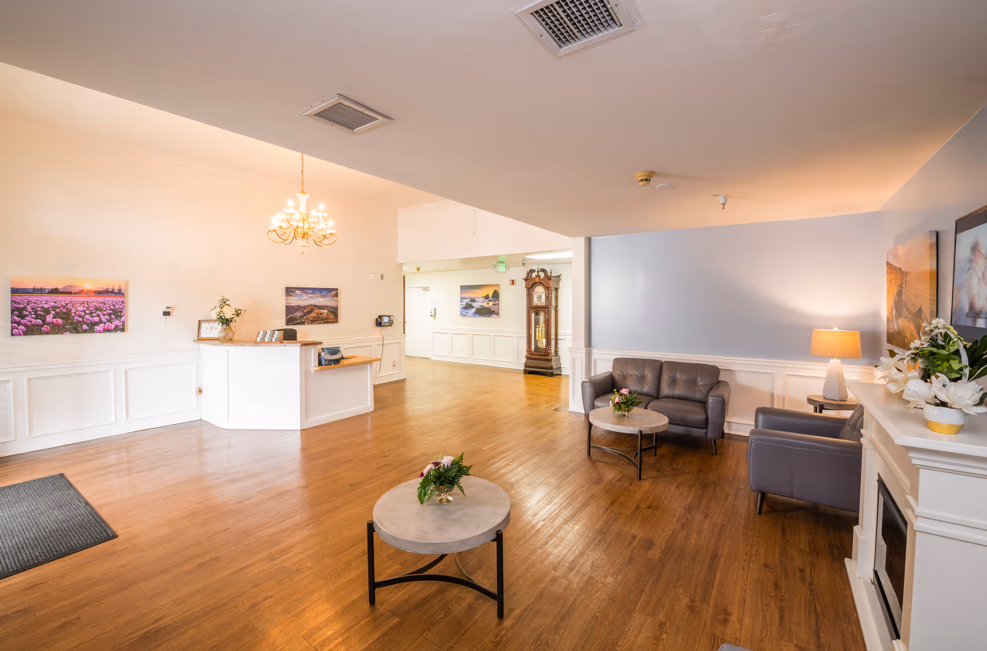A bright and spacious reception area of Anaheim Healthcare Center featuring a wooden floor, a white reception desk with a small plant and framed pictures, a chandelier hanging from the ceiling, a seating area with a gray couch and armchair around a round coffee table with flowers, a grandfather clock in the background, and framed landscape photographs on the walls.