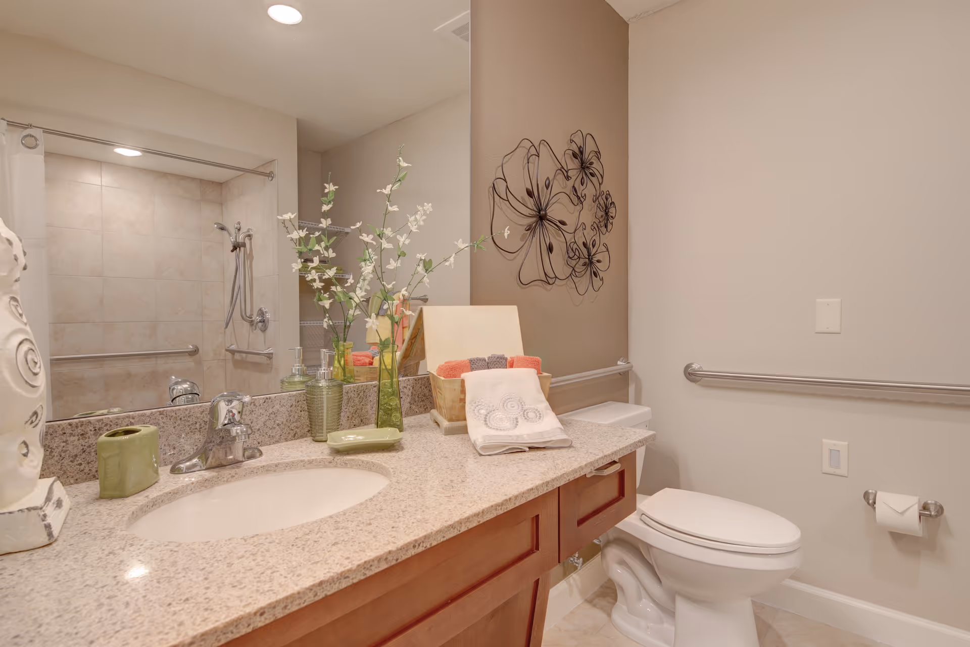 A clean and well-lit bathroom featuring a granite countertop with a sink, decorative flowers in a vase, a soap dish, and neatly folded towels. The bathroom includes a toilet with a grab bar on the wall and a shower area with a handheld showerhead and grab bars, reflecting accessibility features.