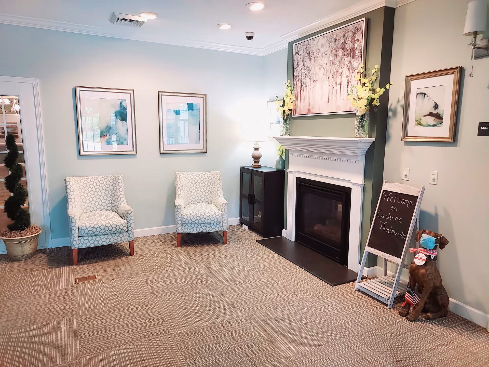 A cozy sitting area in a senior living facility with two patterned armchairs, a fireplace with a mantel decorated with flowers and a large painting above it, a small cabinet with a lamp, framed artwork on the walls, and a small chalkboard sign that reads 'Welcome to Cadence Huntersville' next to a dog statue wearing a face mask.