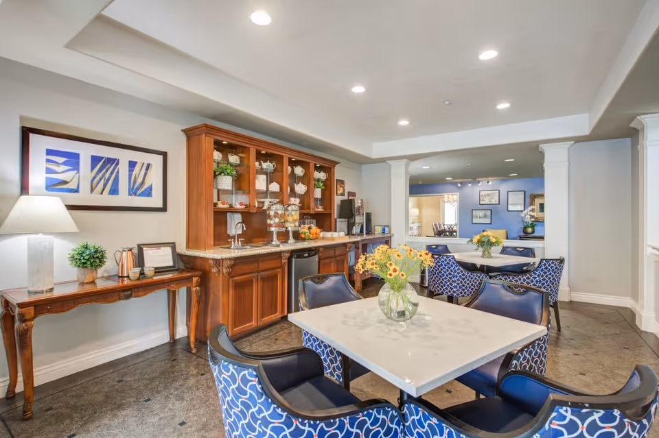 A bright and inviting dining area in a senior living facility featuring a square white table with a vase of yellow flowers, surrounded by blue patterned chairs. In the background, there is a wooden cabinet with glass doors displaying teapots and cups, a countertop with a coffee machine, and a side table with a lamp and framed artwork on the wall.