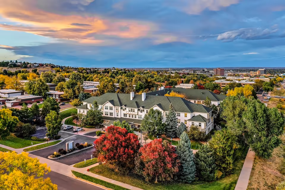 Aerial view of Woodlake Legacy Senior Living facility surrounded by trees with autumn foliage, residential buildings, and a partly cloudy sky at sunset.