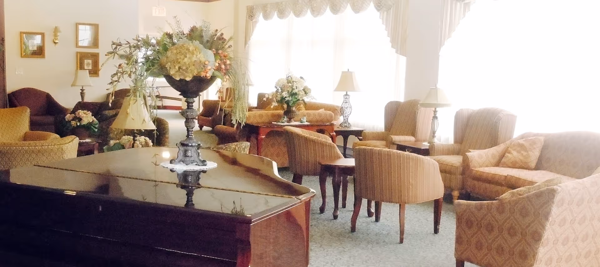A bright and cozy living room with multiple upholstered armchairs and sofas arranged around small wooden tables. A large floral arrangement sits on top of a polished grand piano in the foreground. The room is softly lit by table lamps and natural light coming through large windows with sheer curtains.