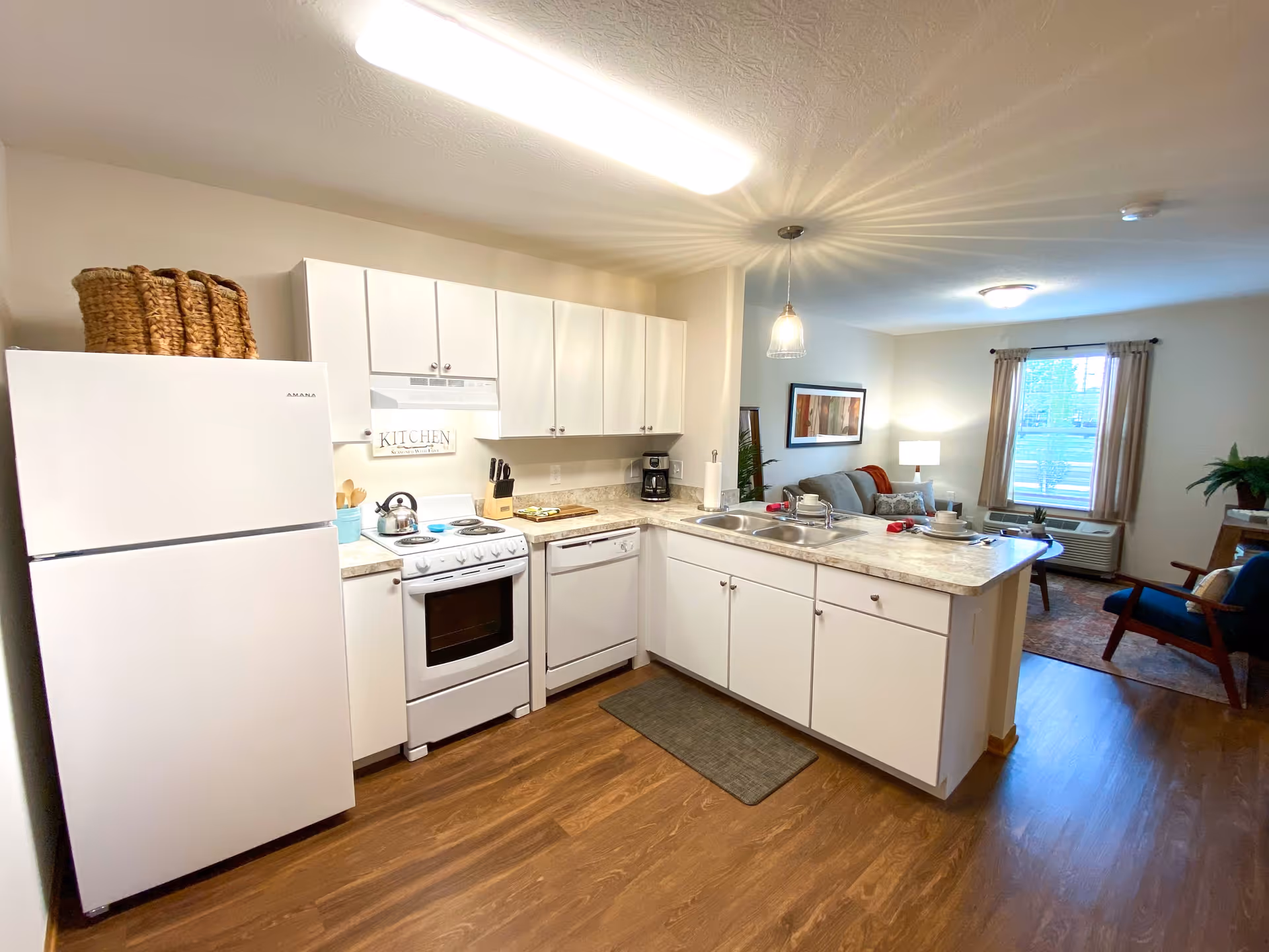 A bright and clean kitchen area with white cabinets, a white refrigerator, stove, and dishwasher. The kitchen has a countertop with a sink and some dishes set on it. Beyond the kitchen is a living room area with a sofa, armchair, window with curtains, and a lamp on a side table. The floor is wooden, and the walls are painted light beige.