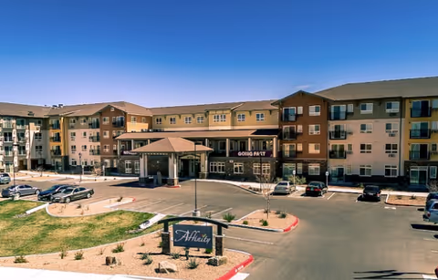 Exterior view of a multi-story senior living facility named Affinity at Albuquerque with a parking lot in front, landscaped areas with grass and desert plants, and a clear blue sky above.