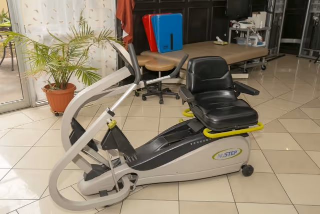 A NuStep exercise machine with a black seat and silver frame is placed on a tiled floor in a rehabilitation center room. In the background, there is a treatment table, a rolling stool, some exercise mats, a potted plant near a glass door, and storage shelves with medical supplies.