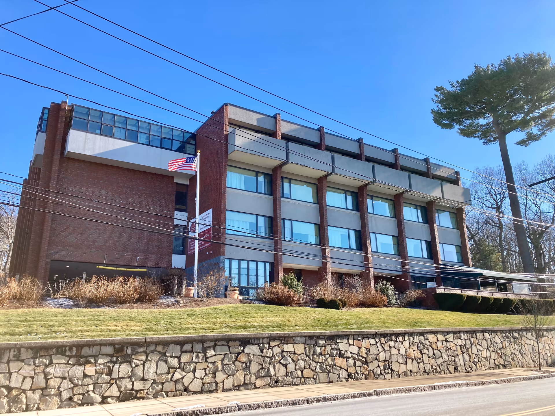 Four-story brick-and-concrete building with rows of windows, an American flag, and a stone retaining wall under a clear blue sky.