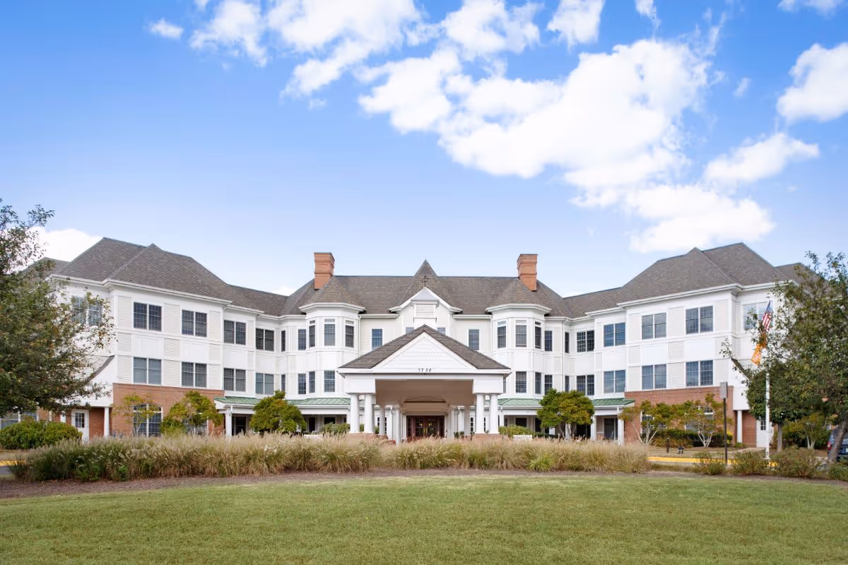 Front exterior view of a large, three-story senior living facility building with white siding and brick accents, multiple windows, a covered entrance with columns, surrounded by landscaped bushes and trees under a blue sky with scattered clouds.