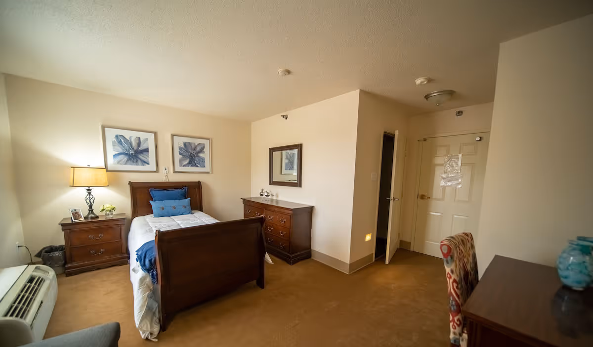 A cozy bedroom in a senior living facility featuring a single wooden bed with white and blue bedding, a wooden nightstand with a lamp and small decor, a wooden dresser with a mirror above it, a patterned chair, and a door leading to another room. The walls are light beige and the carpet is brown.