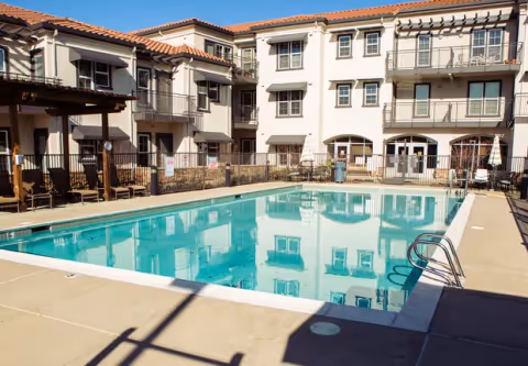Outdoor swimming pool in the courtyard of a multi-story senior living facility with balconies and a pergola-covered seating area on the left side.