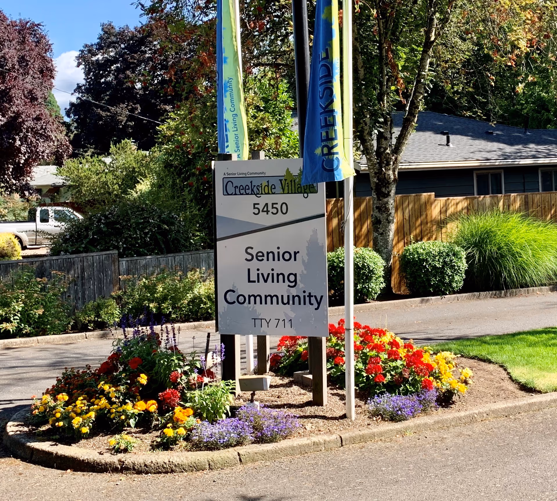 A sign for Creekside Village Senior Living Community at 5450, surrounded by colorful flowers and greenery, with flags and trees in the background.