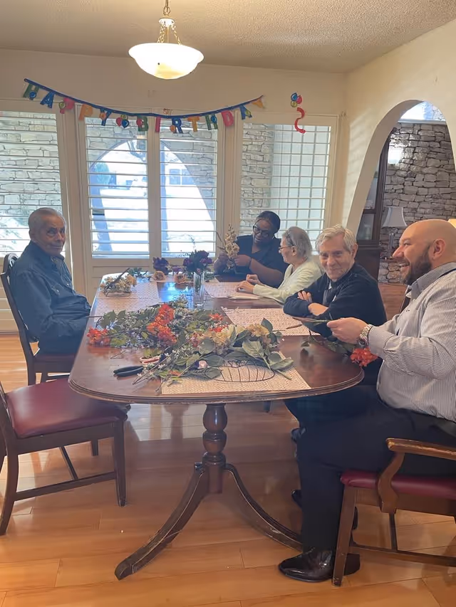 A group of five people sitting around a wooden dining table in a well-lit room with large windows and a Happy Birthday banner hanging above. The table is decorated with various flowers and leaves. The people appear to be engaged in a craft or floral activity, smiling and interacting with each other.