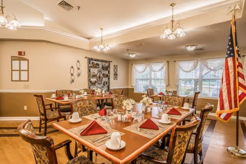 A dining room in a senior living facility with several tables set for a meal. Each table has white cups, silverware, glasses, and red folded napkins. The room has patterned chairs, large windows with sheer curtains, and an American flag standing in the corner. The walls are decorated with a tapestry and wall hangings, and the ceiling has multiple light fixtures.