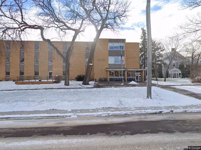 Front exterior view of a three-story brick building with several windows, surrounded by snow-covered ground and leafless trees. There is a flagpole with an American flag near the entrance and a white gazebo to the right side of the building.
