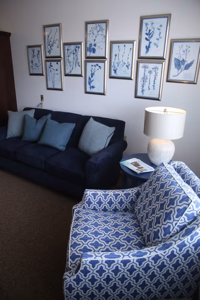 A cozy living room area with a dark blue sofa adorned with multiple blue and gray cushions, a patterned blue and white armchair, a small round side table with a white textured lamp and a magazine on it, and a wall decorated with nine framed botanical prints featuring blue floral illustrations.