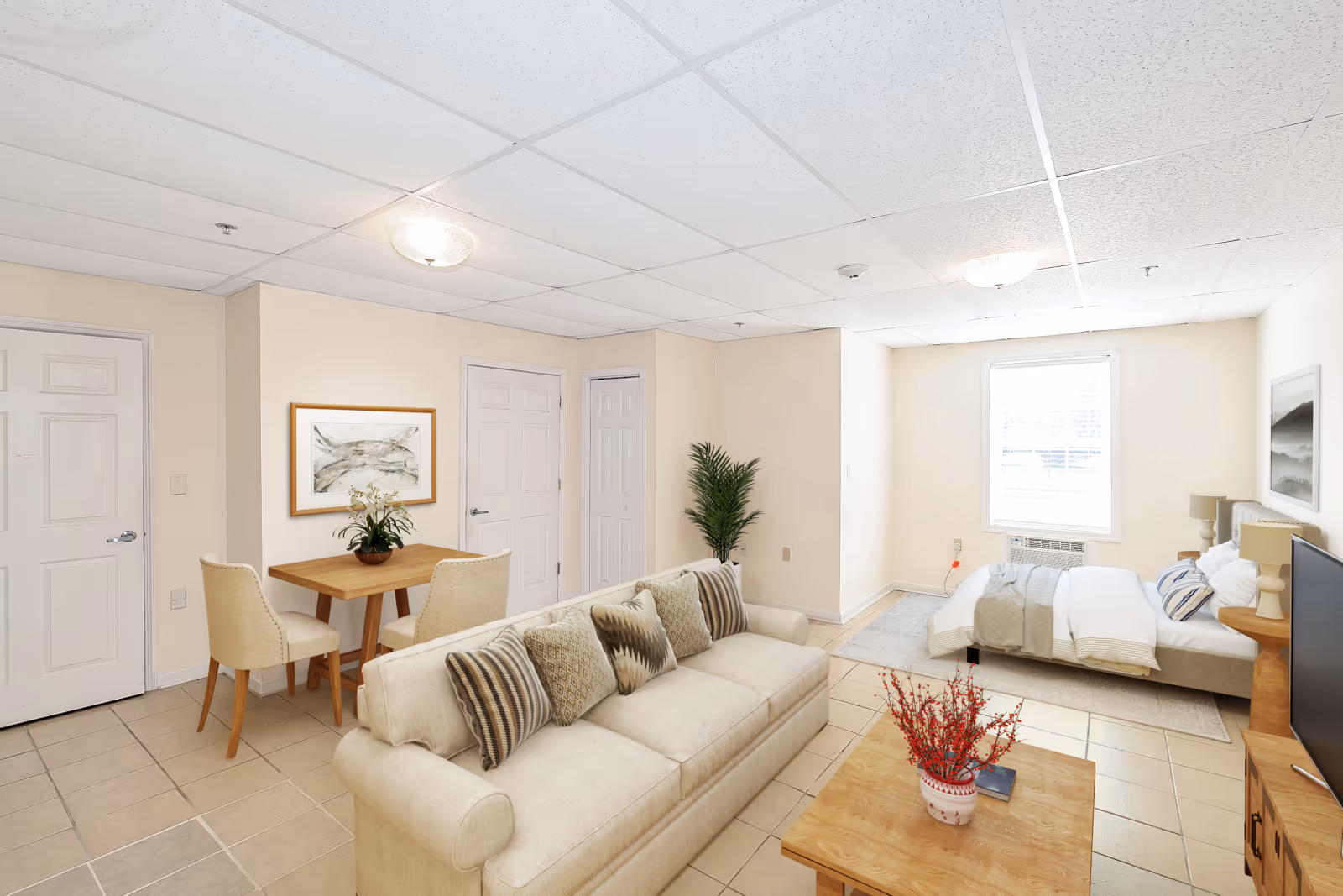 A bright and clean senior living studio apartment featuring a beige sofa with patterned cushions, a wooden coffee table with a vase of red flowers, a small dining table with two upholstered chairs, a bed with white and beige bedding, a window with blinds, and light-colored tiled flooring. The walls are painted cream, and there are two framed artworks and a potted plant in the corner.