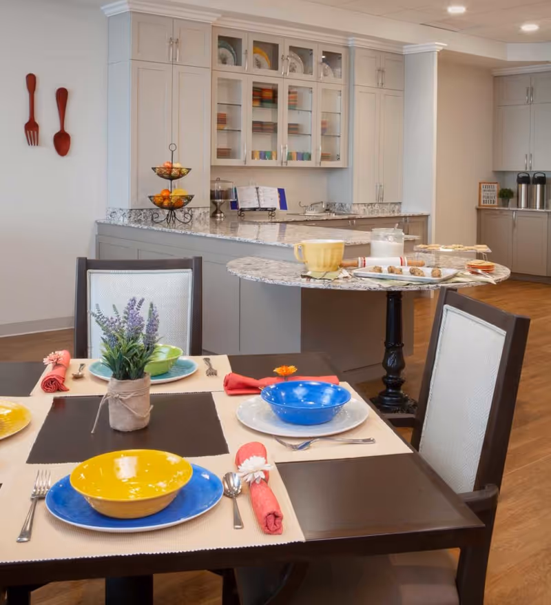 A bright dining area with a set table of colorful bowls and napkins and a marble-topped kitchen island with cabinets and serving dishes in the background.