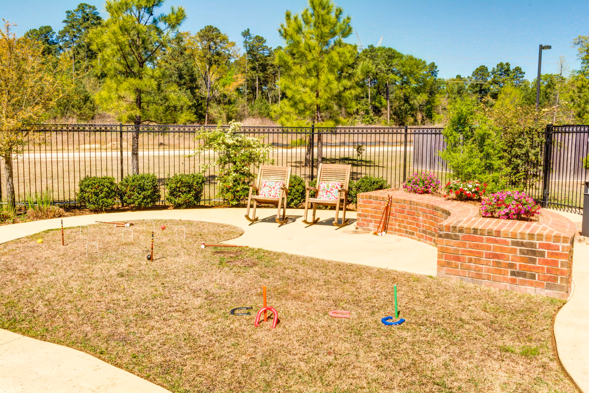 Sunlit fenced courtyard with lawn games, two wooden chairs, and a curved brick planter filled with flowers.