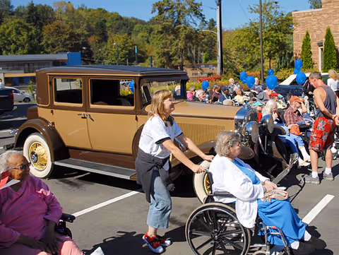 An outdoor gathering at Washington Care Center with elderly people, some in wheelchairs, and a woman pushing a wheelchair. A vintage brown car is parked nearby, and blue balloons are visible in the background with trees and a building under a clear sky.