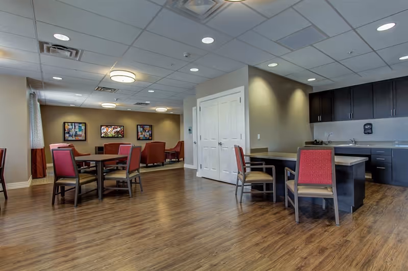Interior view of a senior living facility common area with wooden flooring, several tables and chairs with red upholstery, a kitchenette with dark cabinets and a sink, and framed artwork on the far wall.