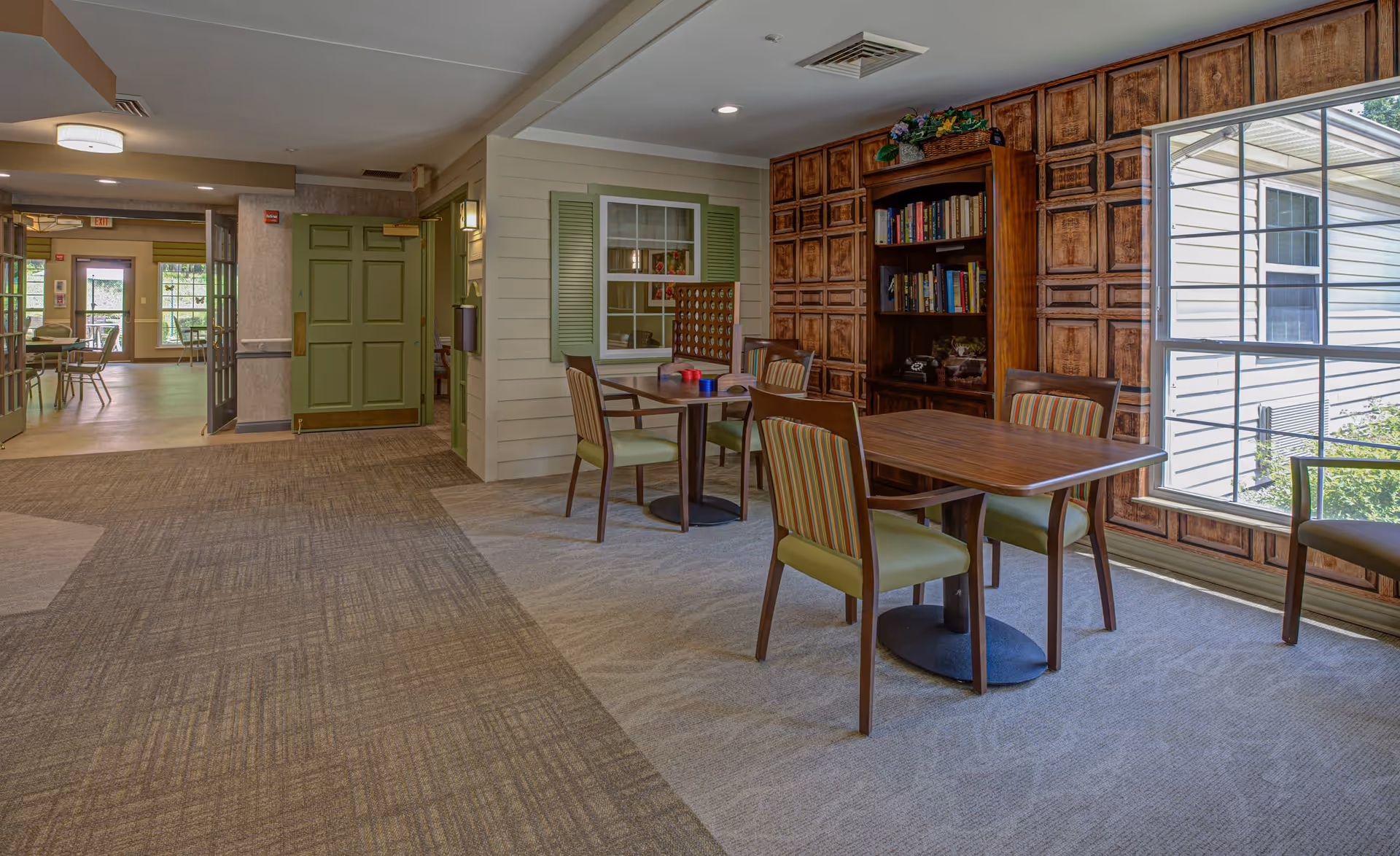 A cozy interior common area with two wooden tables and several chairs with green and striped upholstery. There is a large window letting in natural light, a wooden bookshelf filled with books, and a decorative wooden paneled wall. The floor is carpeted and there is a green door and another room visible in the background.