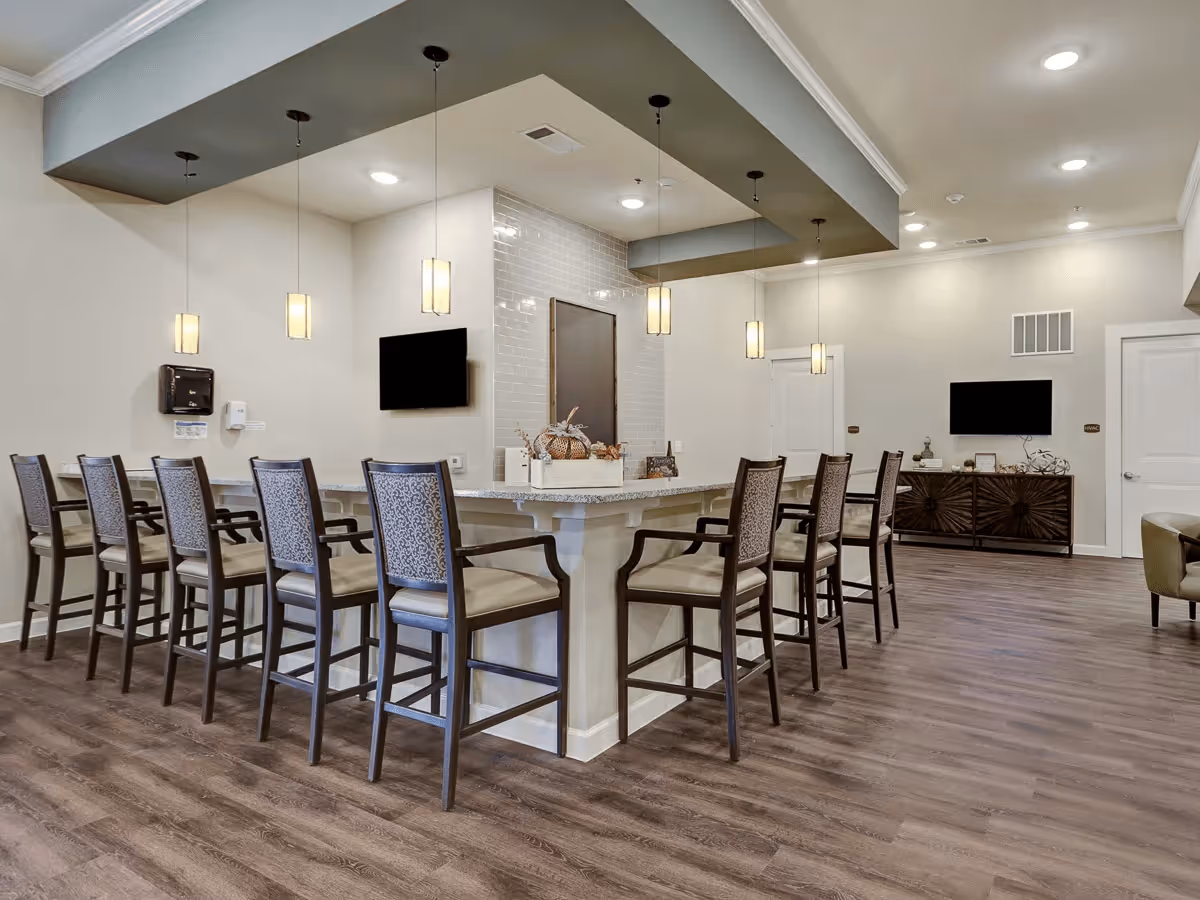 Interior view of a communal dining area with a long counter surrounded by high chairs. The room features pendant lights hanging from the ceiling, a wall-mounted TV, and decorative items on the counter. The flooring is wood-style, and the walls are painted in neutral tones.