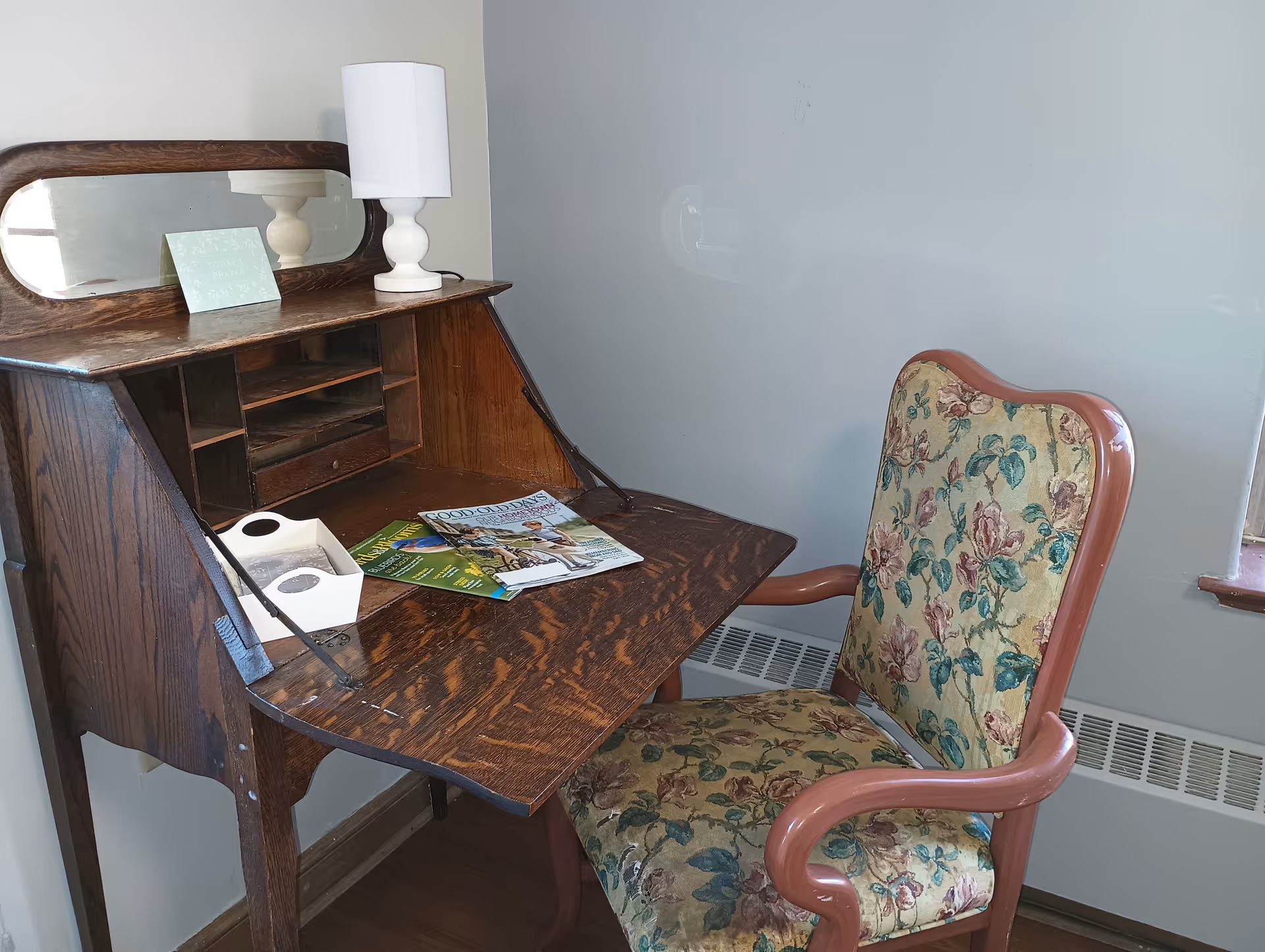 A vintage wooden writing desk with a fold-down surface, a small white lamp, a card that says 'Today's Prayer', and some magazines on the desk. Next to the desk is an upholstered chair with a floral pattern and wooden armrests. The setting appears to be a quiet corner of a room with light-colored walls and a window.