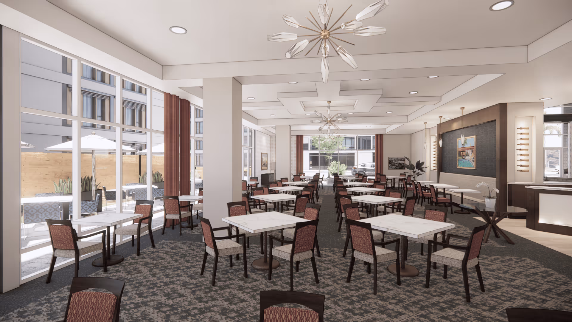 A spacious dining room with multiple square tables and chairs arranged in rows. The room features large windows on one side allowing natural light to enter, with an outdoor patio visible through the windows. The ceiling has modern light fixtures and recessed lighting. The walls are decorated with framed artwork and plants, and there is a small counter area on the right side.