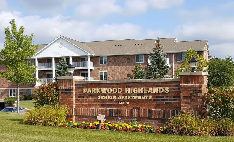 Brick entrance sign reading "Parkwood Highlands Senior Apartments" with flowers and landscaping in front of a multi-story brick apartment building.
