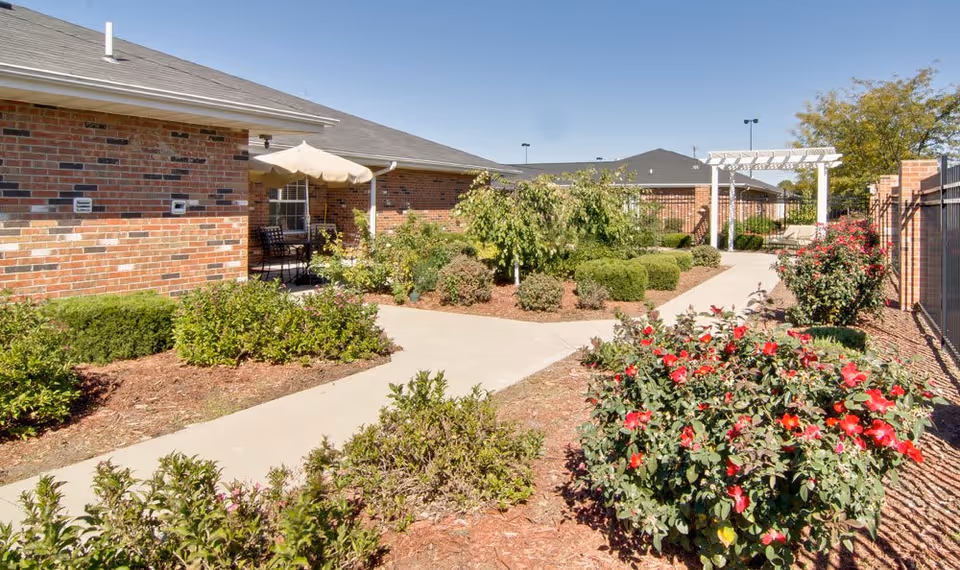 Sunlit courtyard with paved walkways, landscaped beds and red flowering shrubs beside a brick senior living building and a white pergola.