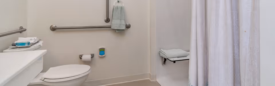 A clean, accessible bathroom featuring a white toilet with a closed lid, grab bars on the walls for support, a towel hanging on one of the bars, a shelf with folded towels and toiletries, and a shower area with a fold-down seat and a white patterned shower curtain.