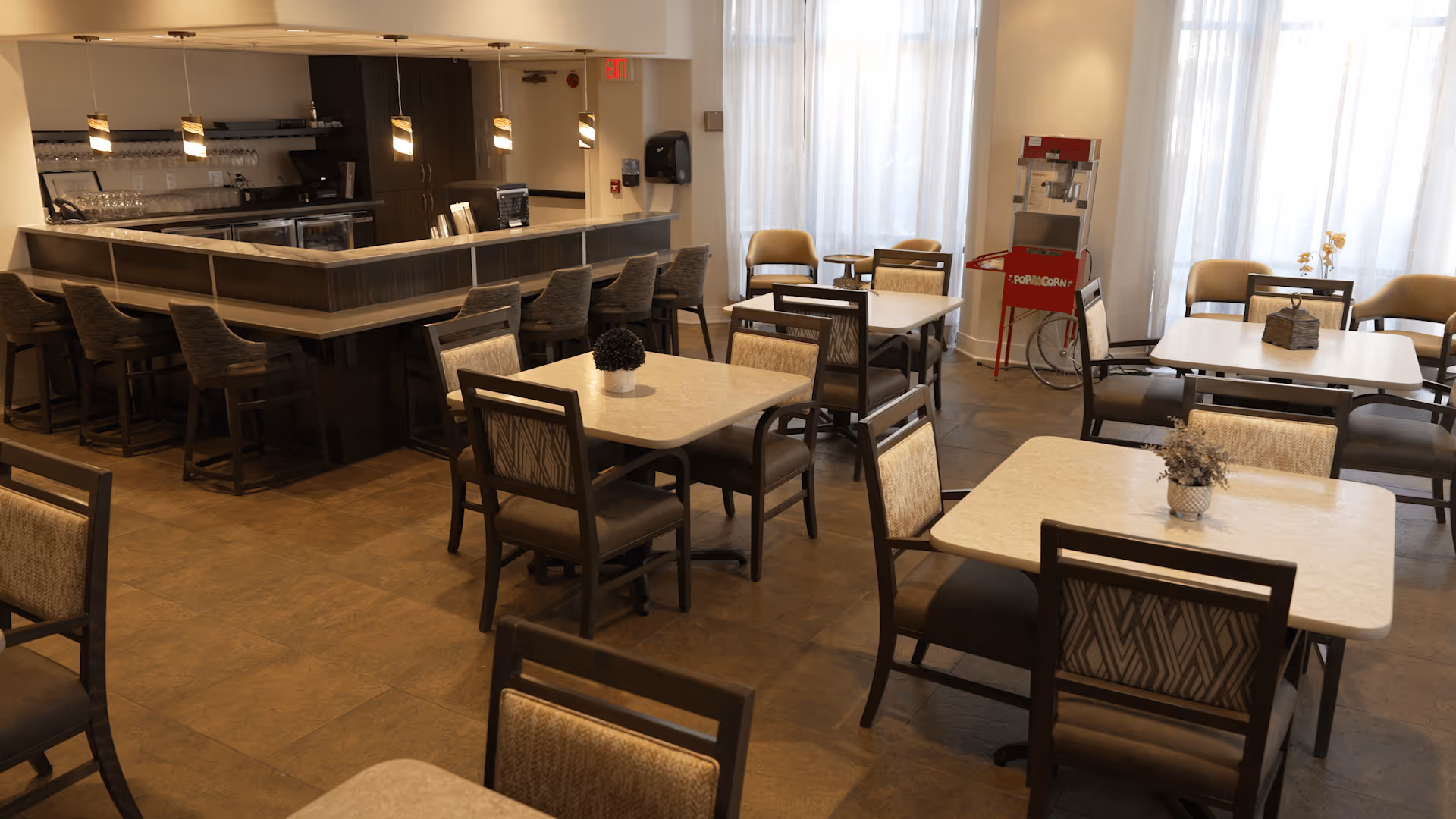 A dining area with several square tables and cushioned chairs arranged neatly. In the background, there is a counter with bar stools and hanging pendant lights above. A red popcorn machine is visible near large windows with sheer white curtains, allowing natural light to fill the room.