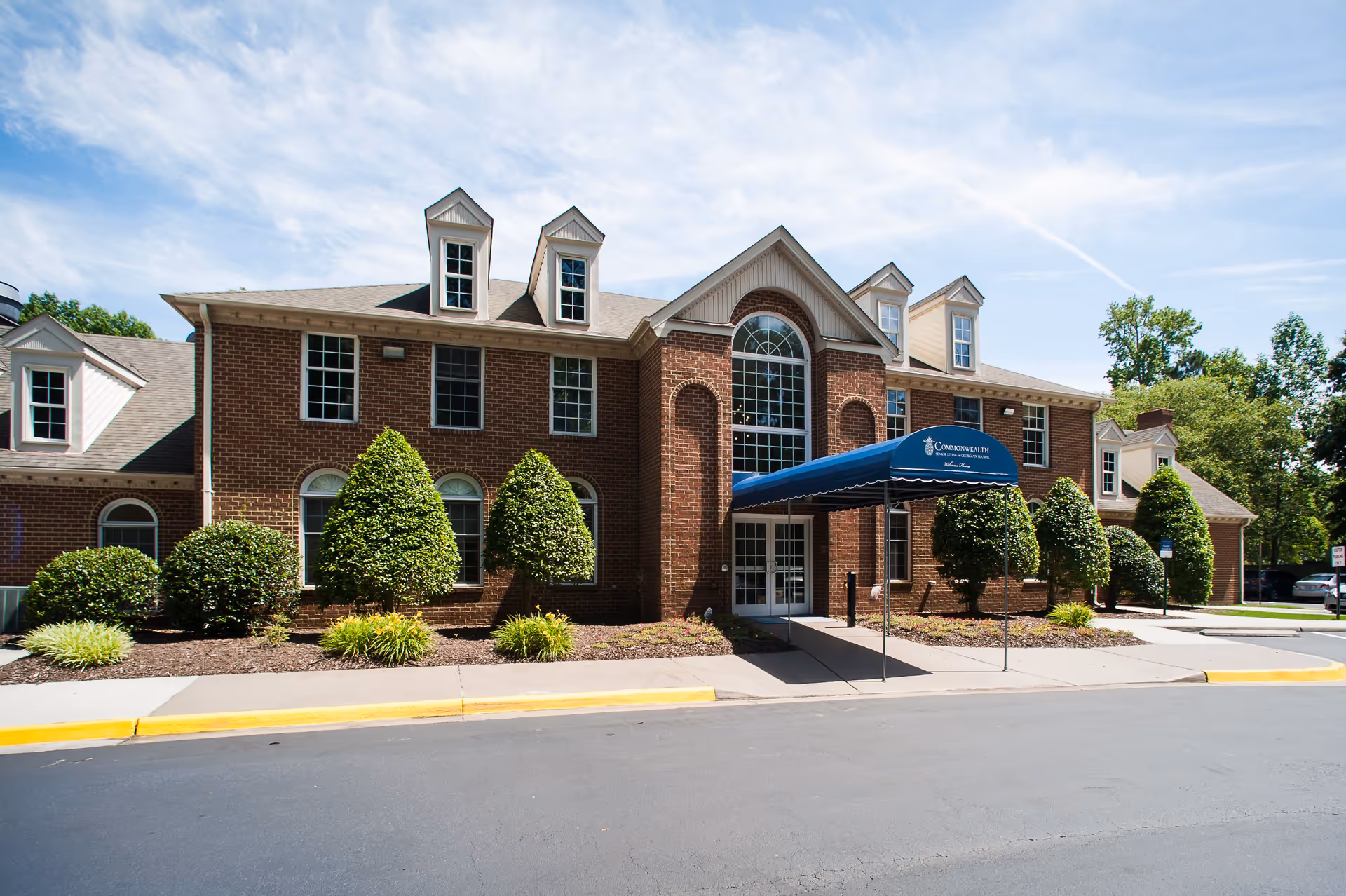 Front exterior view of a two-story brick building with multiple windows and dormers on the roof. There is a blue canopy over the entrance with the text 'Commonwealth Senior Living at Georgian Manor' and 'Welcome Home'. The building is surrounded by neatly trimmed bushes and small trees, with a clear blue sky above.