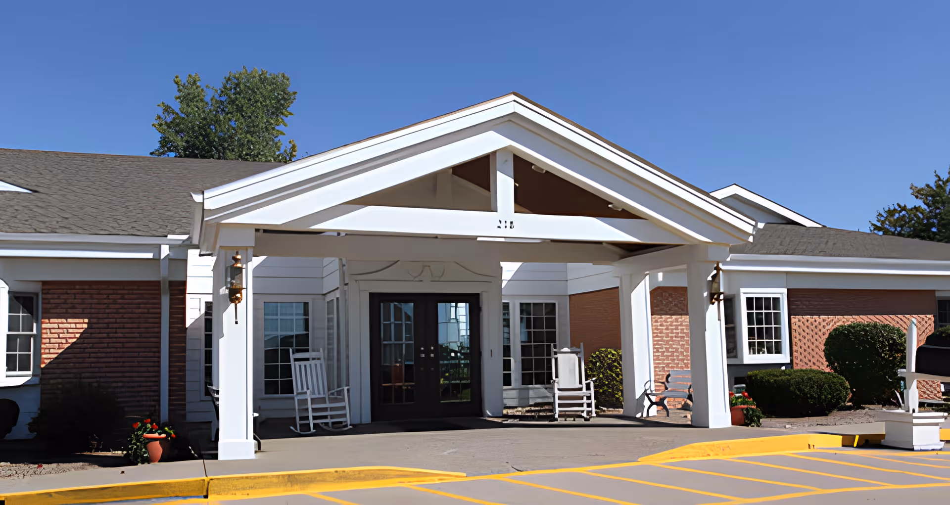 Entrance of Shelbina Villa Lifecare facility showing a covered drop-off area with white pillars and rocking chairs on either side of the double glass doors. The building exterior features red brick and white siding with windows and landscaping including bushes and potted plants. The sky is clear and blue.