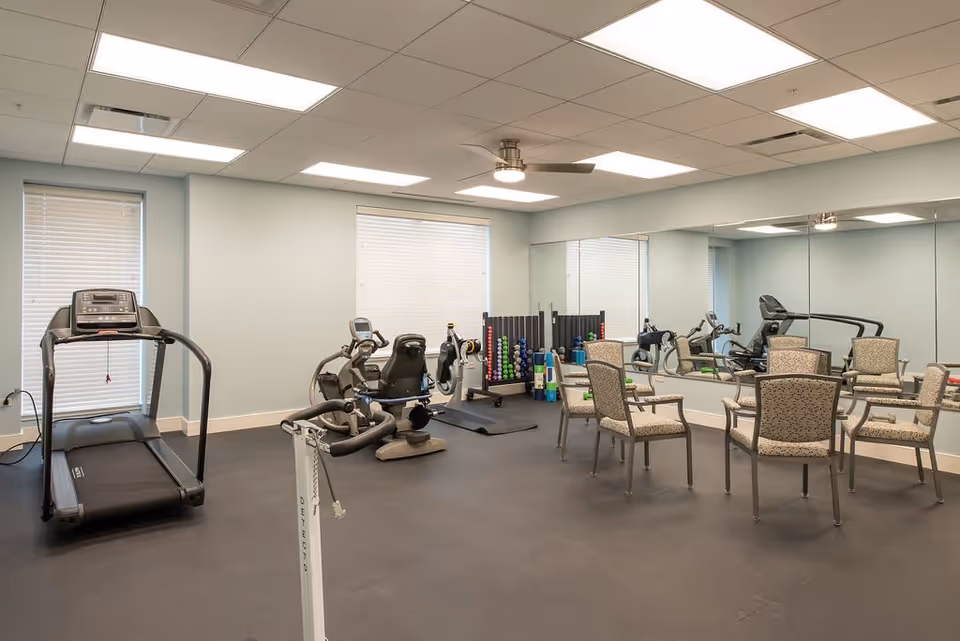 A fitness room with exercise equipment including a treadmill, stationary bikes, and a rack of colorful dumbbells and exercise mats. There are six patterned chairs arranged in a circle in the middle of the room. The walls are light blue with two windows covered by blinds and a large mirror covering one wall. The ceiling has recessed lighting and a ceiling fan.