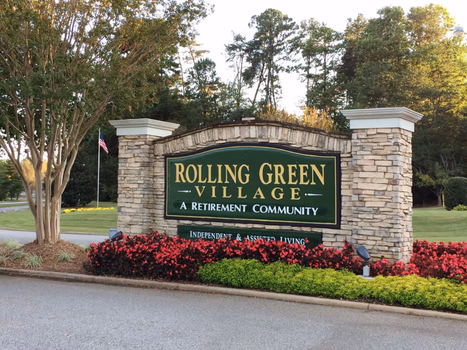 Stone entrance sign for Rolling Green Village, a retirement community, surrounded by red and green landscaping plants with trees and an American flag in the background.