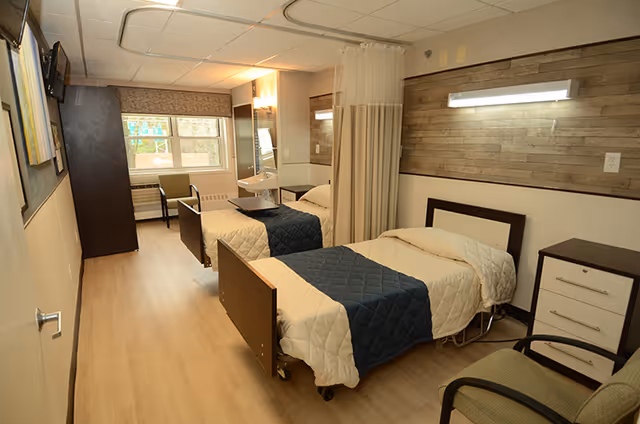 A clean and well-lit nursing facility room with two single beds separated by a curtain. Each bed has a beige quilt and a dark blue blanket. The room features light wood flooring, a window with a view outside, a chair near the window, a small bedside table with drawers, and a wall-mounted light above each bed. There is a sink and mirror on the far wall, and the walls have a wood panel accent behind the beds.
