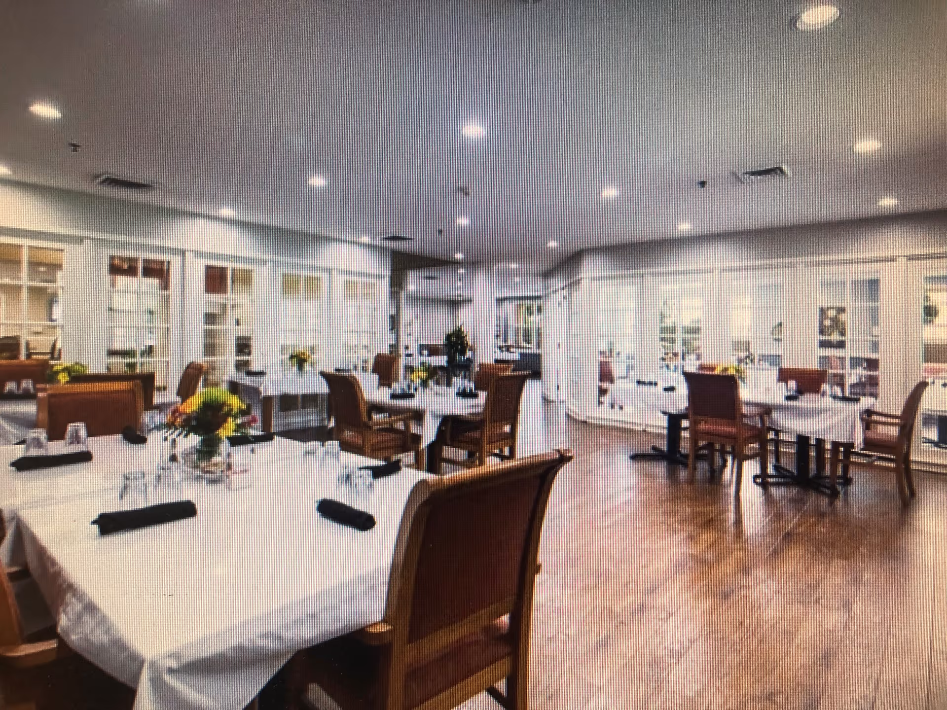 A spacious dining room with several tables covered in white tablecloths, each set with glasses and black napkins. Wooden chairs with red cushions surround the tables. The room has wooden flooring, recessed ceiling lights, and large windows along the walls allowing natural light to enter.