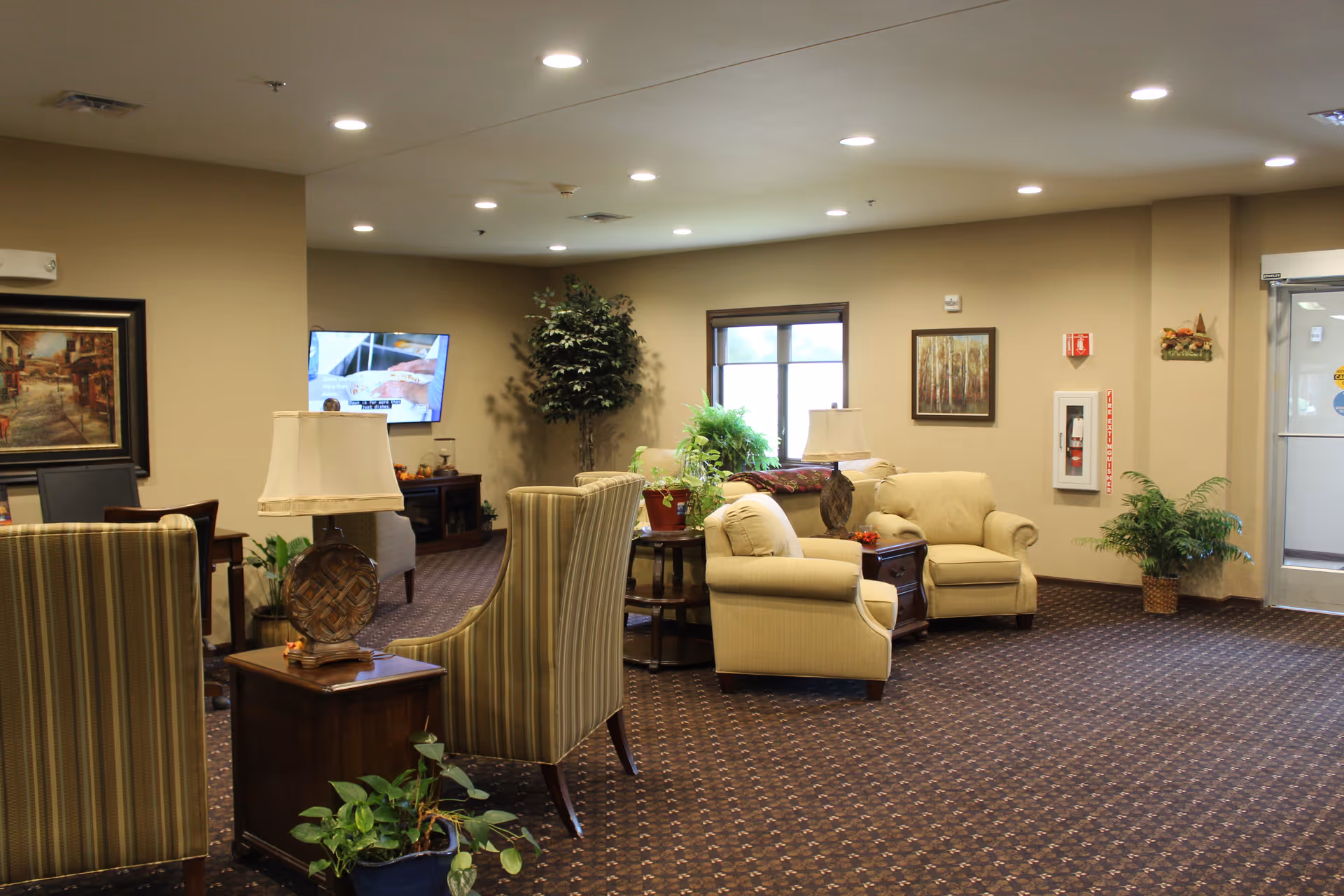 A cozy living room area in a senior living facility with beige armchairs, striped chairs, side tables with lamps, potted plants, framed artwork on the walls, a television mounted on the wall, and a window letting in natural light.