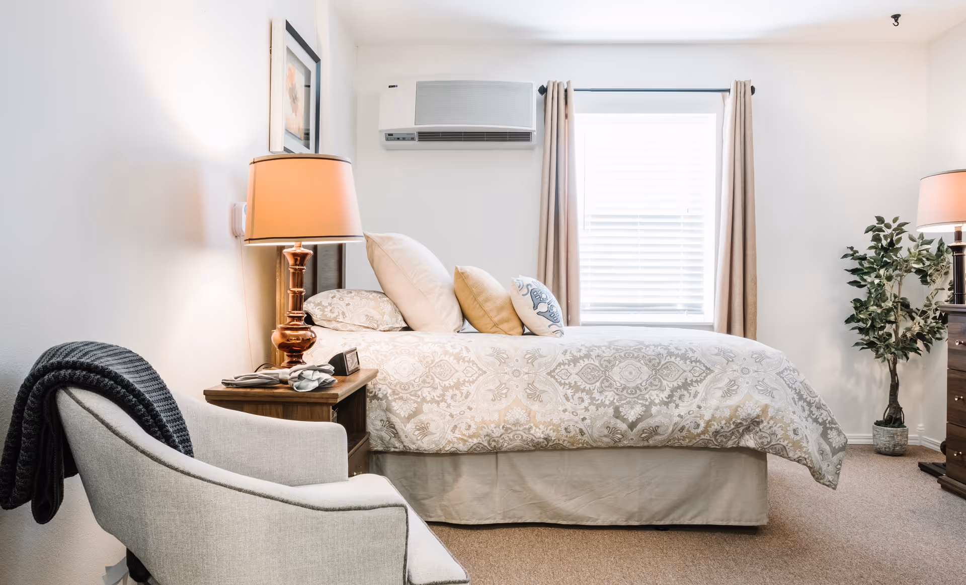 A bright, softly lit bedroom with a made bed, nightstands and lamps, an armchair, and a window with curtains.