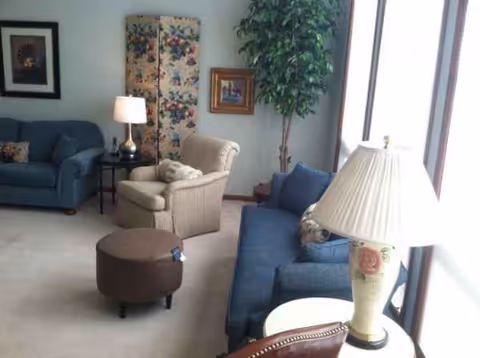 Bright senior living lounge with blue sofa, beige recliner, ottoman, lamp, plant, by tall windows.