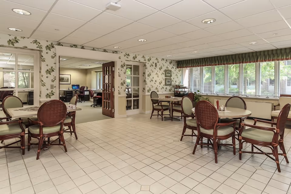 A bright room with several round tables and chairs arranged for dining or socializing. The walls have floral wallpaper and large windows with blinds letting in natural light. An adjoining office space with desks and chairs is visible through a doorway.
