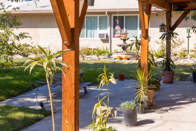 Outdoor patio area with wooden beams supporting a roof, several potted plants on the concrete floor, a bench, and a bird feeder hanging in the background near a building with windows and an air conditioning unit.