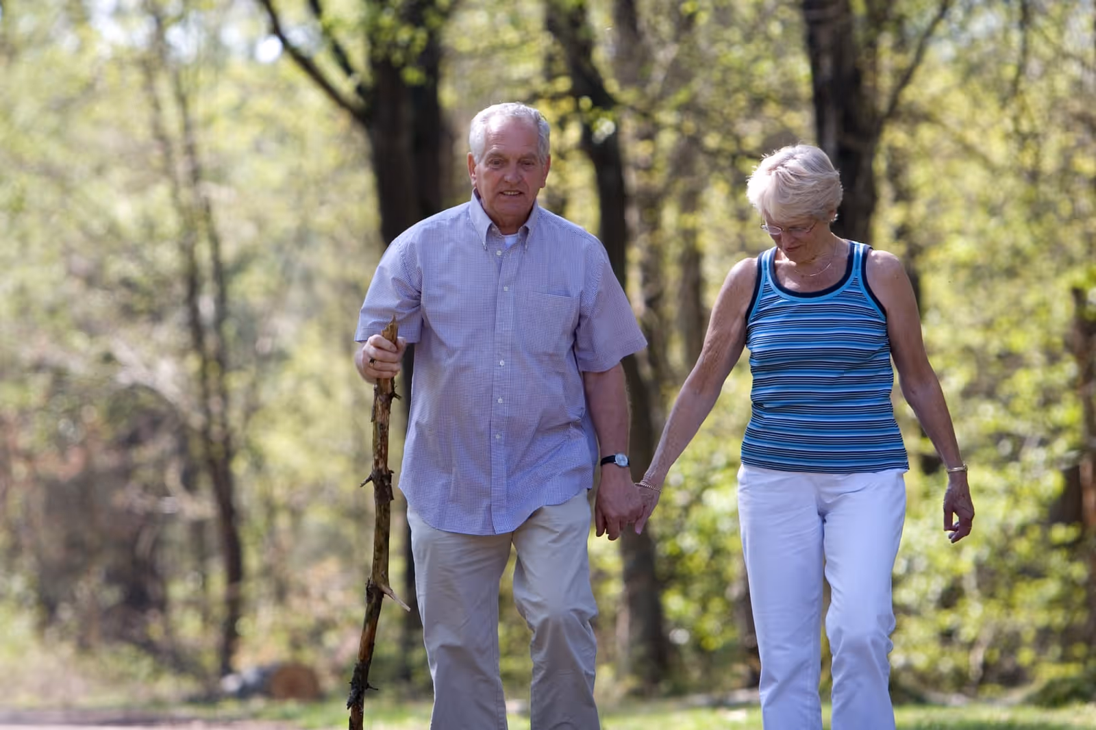 An elderly couple walking hand in hand outdoors on a wooded path, with the man holding a walking stick and both dressed casually in light clothing.