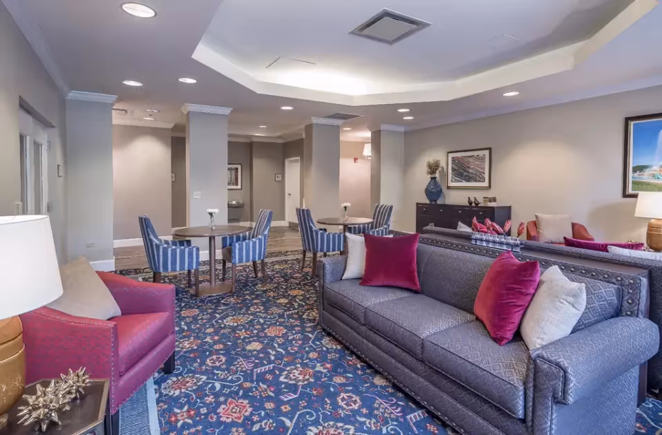 A bright common lounge in a senior living facility with sofas, armchairs, striped dining chairs, small round tables, and decorative pillows.