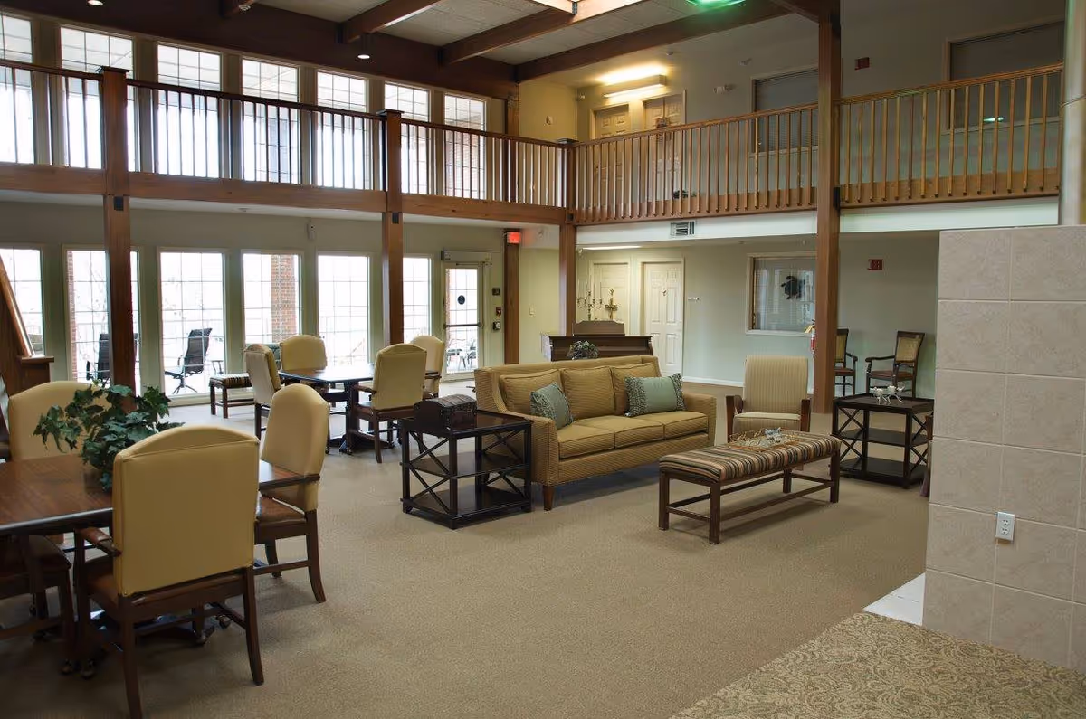 Spacious and well-lit common area in a retirement community featuring multiple seating arrangements including cushioned chairs around tables, a sofa with throw pillows, a striped bench, and side tables. The room has large windows and glass doors letting in natural light, a second-floor balcony with wooden railings, and neutral-colored walls and carpet.