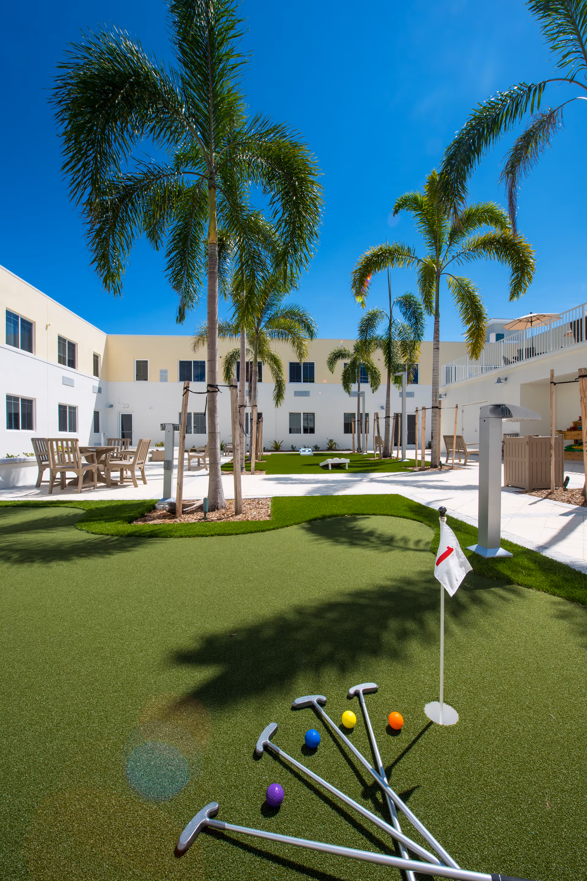 Outdoor courtyard area at The Cabana at Jensen Dunes featuring a putting green with colorful golf balls and putters, surrounded by palm trees, patio tables and chairs, and a clear blue sky.